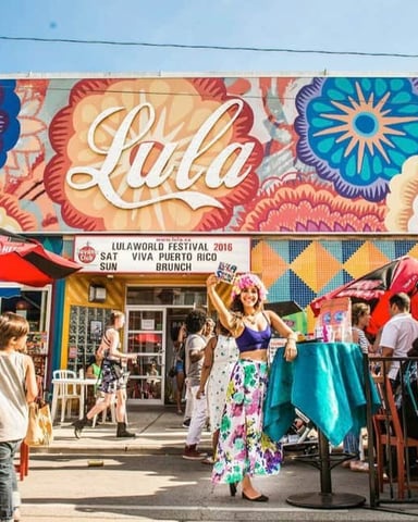 Colorful street festival with large mural featuring flowers and "Lala" text, outdoor seating, red umbrellas, and crowds of people