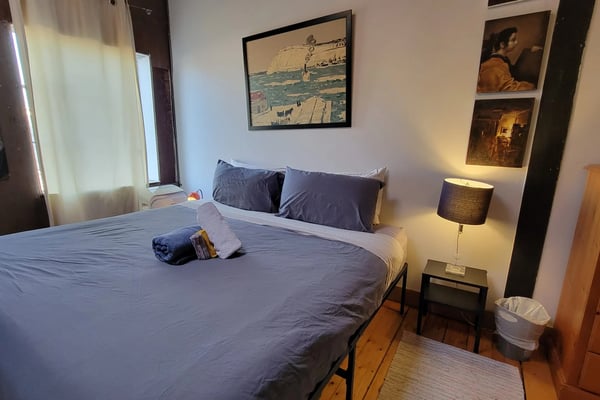 Bedroom with queen bed, blue bedding, nightstand with lamp, framed artwork on walls, wooden dresser, and window with curtains.