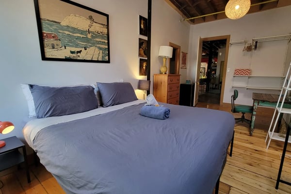 Bedroom with queen bed, blue bedding, wooden floors, exposed ceiling beams, framed artwork, and open shelving with decorative items