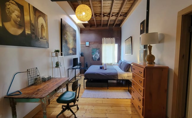 Bedroom with queen bed, wooden desk, dresser, exposed beam ceiling, and portrait artwork on walls